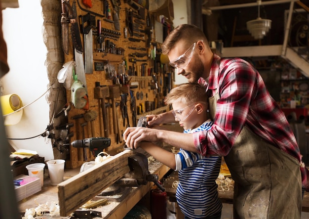 concepto de familia, carpintería, carpintería y personas - padre e hijo pequeño con avión trabajando con tablones de madera en el taller