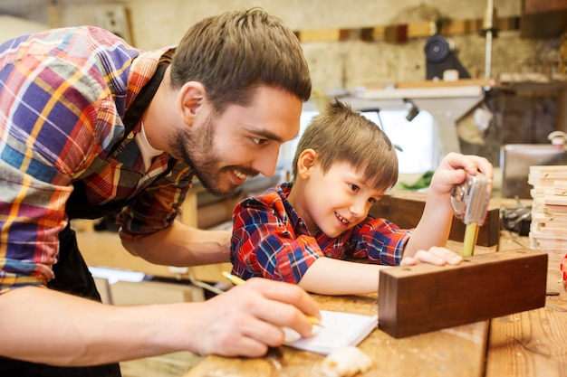 concepto de familia, carpintería, carpintería y personas - padre e hijo pequeño con regla y lápiz midiendo tablones de madera y escribiendo dimensiones en un cuaderno en el taller