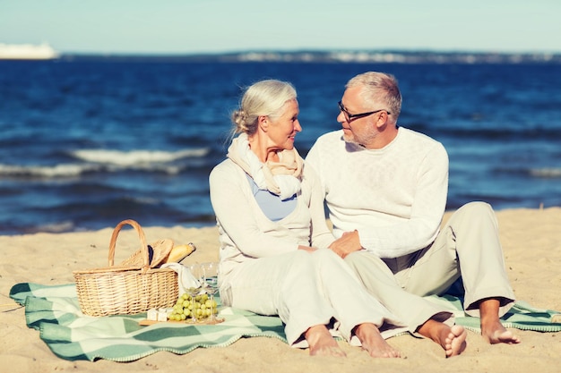 Foto concepto de familia, edad, vacaciones, ocio y personas - feliz pareja mayor con cesta de picnic sentada en una manta en la playa de verano