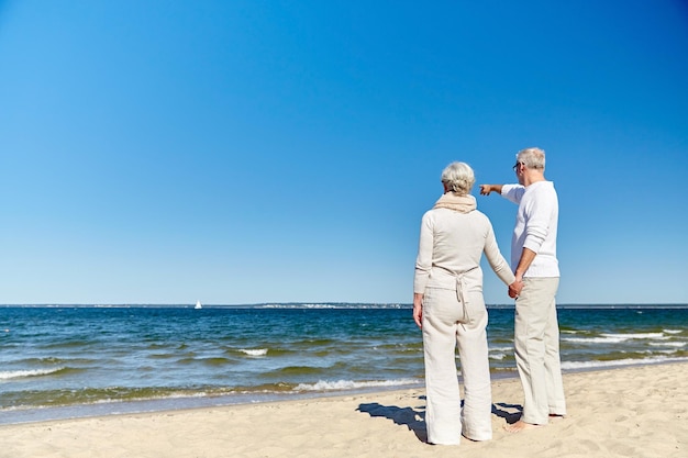 concepto de familia, edad, viajes, turismo y personas - feliz pareja de ancianos señalando con el dedo algo en la playa de verano