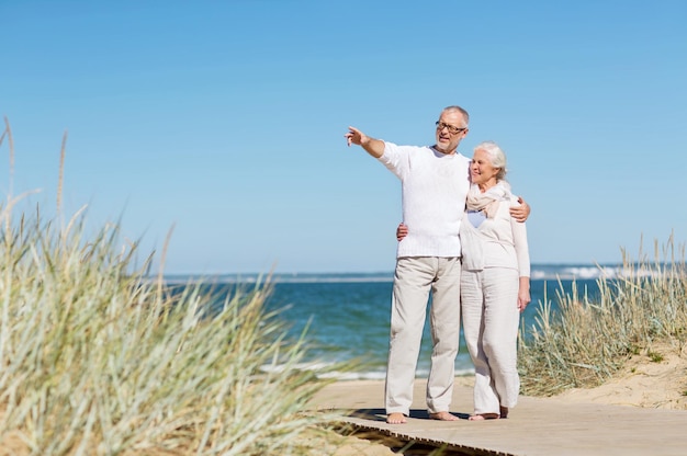 concepto de familia, edad, viajes, turismo y personas - feliz pareja mayor abrazándose y señalando con el dedo en la playa de verano