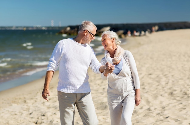 concepto de familia, edad, viajes, turismo y personas - feliz pareja mayor tomándose de la mano y caminando en la playa de verano