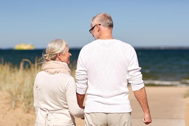 concepto de familia, edad, viajes, turismo y personas - feliz pareja mayor tomándose de la mano y caminando en la playa de verano