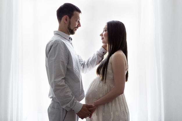 Concepto de familia, embarazo y paternidad - retrato de una feliz pareja embarazada frente a la ventana en casa