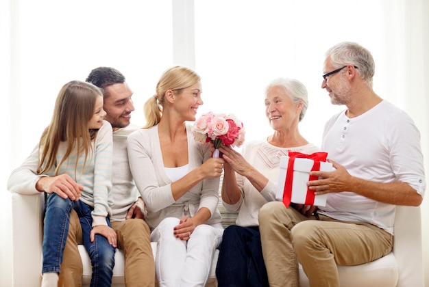 concepto de familia, felicidad, generación, vacaciones y personas - familia feliz con ramo de flores y caja de regalo sentada en el sofá en casa