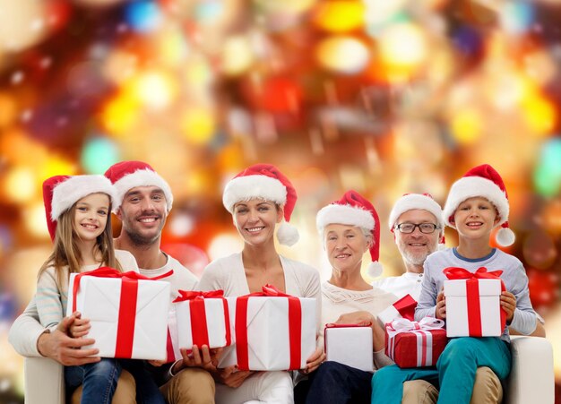 concepto de familia, felicidad, generación, vacaciones y personas - familia feliz con sombreros de ayudante de santa con cajas de regalo sentadas en el sofá sobre fondo de luces rojas