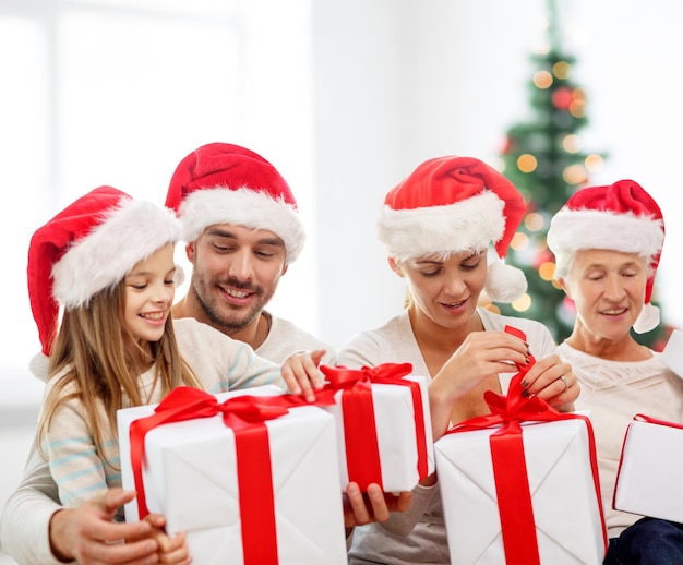 concepto de familia, felicidad, generación, vacaciones y personas - familia feliz con sombreros de ayudante de santa con cajas de regalo sentadas en el sofá sobre la sala de estar y el fondo del árbol de Navidad