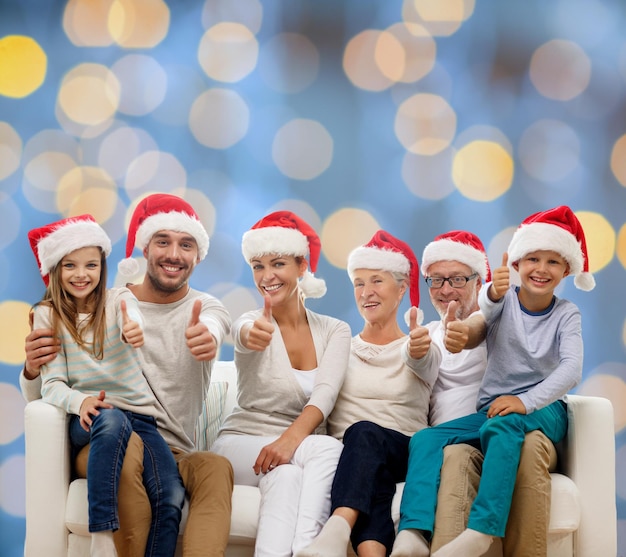 concepto de familia, felicidad, generación, vacaciones y personas - familia feliz con sombreros de ayudante de santa sentados en el sofá y mostrando gestos de aprobación sobre fondo de luces azules