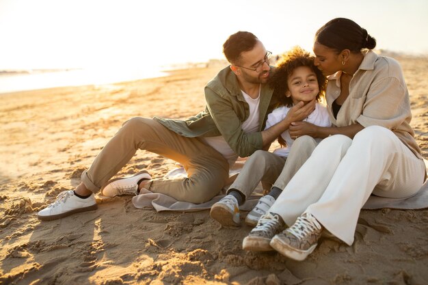 Concepto de familia feliz padres multirraciales y su hijo sentados en la playa disfrutando y gastando