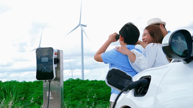 Concepto de familia feliz progresista disfrutando de su tiempo en un parque eólico con un vehículo eléctrico Vehículo eléctrico impulsado por energía limpia renovable de un generador de turbina eólica para una estación de carga