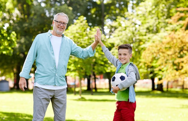 concepto de familia, generación, gesto, deporte y personas - feliz abuelo y nieto con pelota de fútbol haciendo cinco en el parque de verano