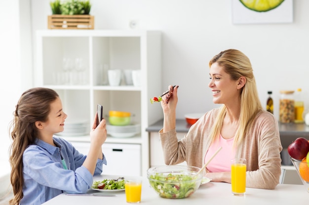concepto de familia y gente - niña feliz con teléfono inteligente cenando y fotografiando a su madre en la cocina de casa