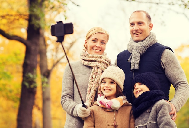 concepto de familia, infancia, temporada, tecnología y personas - familia feliz fotografiando con smartphone y selfie stick en el parque de otoño