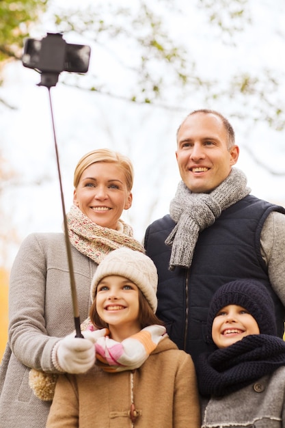 concepto de familia, infancia, temporada, tecnología y personas - familia feliz tomando selfie con smartphone y monopod en el parque de otoño