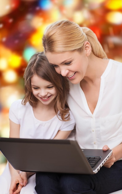 concepto de familia, infancia, vacaciones, tecnología y personas - madre sonriente y niña pequeña con computadora portátil sobre fondo de luces rojas