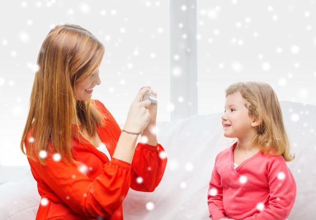 concepto de familia, infancia, vacaciones, tecnología y personas - madre sonriente tomando fotos de su hija con una cámara digital en casa