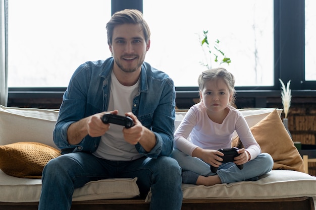 Concepto de familia, juegos y entretenimiento: padre feliz e hija pequeña con gamepads jugando videojuegos en casa.