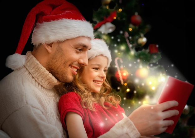 concepto de familia, navidad, navidad, invierno, felicidad y personas - padre e hija sonrientes en el libro de lectura de sombreros de ayudante de santa