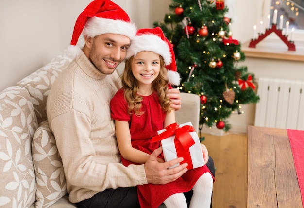 concepto de familia, navidad, navidad, invierno, felicidad y personas - padre e hija sonrientes en sombreros de ayudante de santa con caja de regalo
