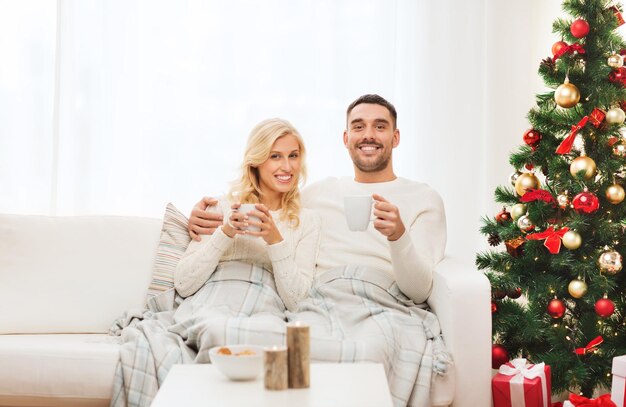 concepto de familia, Navidad, vacaciones, amor y personas - pareja feliz cubierta de té a cuadros bebiendo té y sentada en el sofá en casa