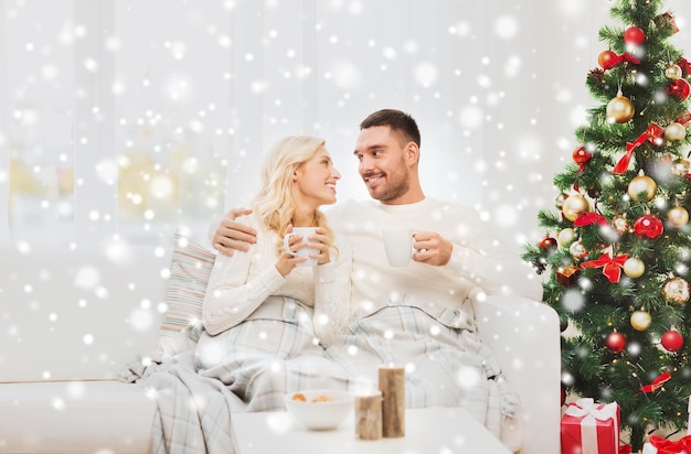 concepto de familia, Navidad, vacaciones, amor y personas - pareja feliz cubierta de té a cuadros bebiendo té y sentada en el sofá en casa