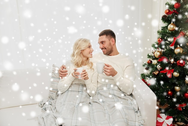 concepto de familia, Navidad, vacaciones, amor y personas - pareja feliz cubierta de té a cuadros bebiendo té y sentada en el sofá en casa