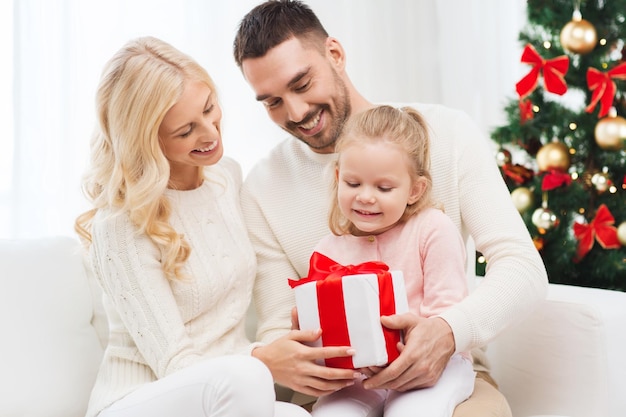 concepto de familia, navidad, vacaciones y personas - madre feliz, padre e hija pequeña con caja de regalo sentada en el sofá en casa