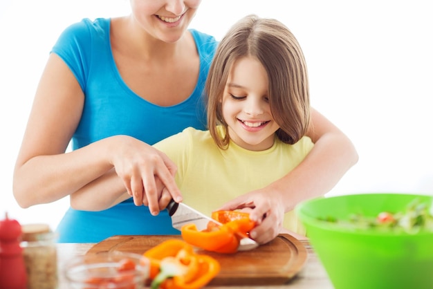 concepto de familia, niño, cocina y hogar - niña sonriente con madre cortando pimienta en la cocina