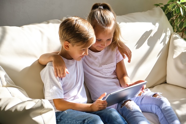 Concepto de familia, niños, tecnología y hogar: hermano y hermana sonrientes con tablet pc en el sofá de la sala de estar