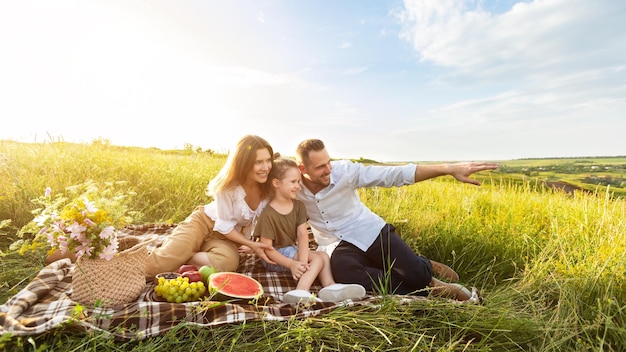 Concepto de familia, ocio y personas. Familiares felices sentados en una manta, padre mostrando un hermoso paisaje