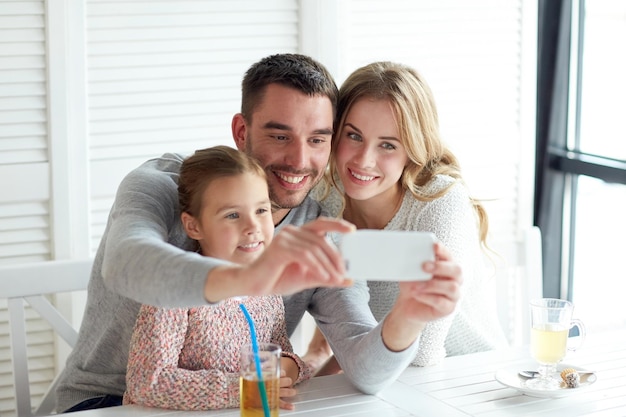 concepto de familia, paternidad, tecnología y personas - feliz madre, padre y niña cenando y tomando selfie por teléfono inteligente en el restaurante