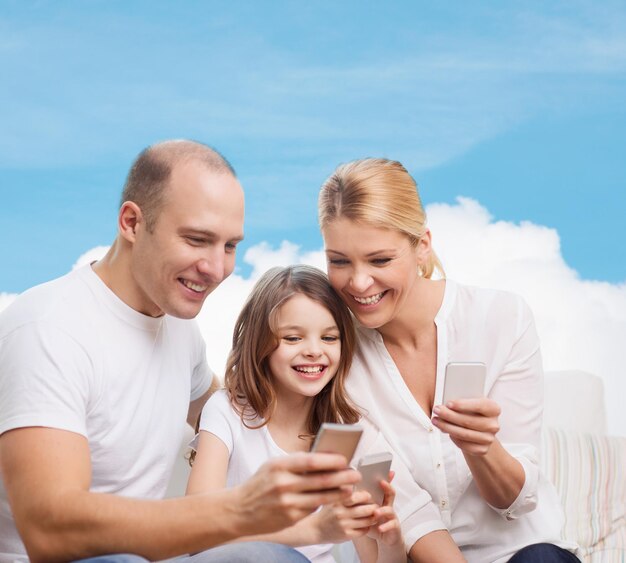 concepto de familia, tecnología y personas: madre, padre y niña sonrientes con smartphones sobre cielo azul y fondo de nube blanca