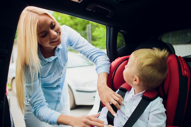 Concepto de familia, transporte, viaje por carretera y personas: mujer feliz que sujeta al niño con el cinturón de seguridad en el automóvil