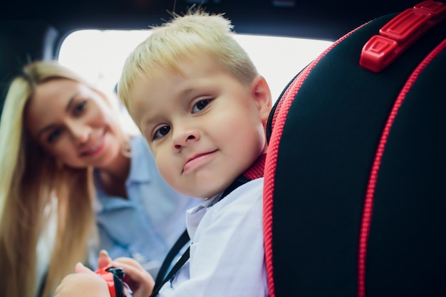 Concepto de familia, transporte, viaje por carretera y personas: mujer feliz que sujeta al niño con el cinturón de seguridad en el automóvil