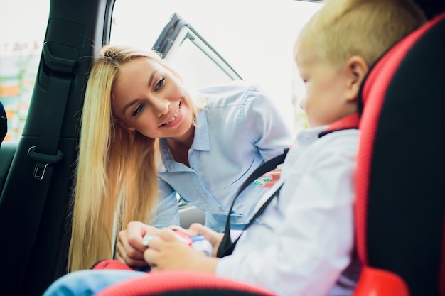 Concepto de familia, transporte, viaje por carretera y personas: mujer feliz que sujeta al niño con el cinturón de seguridad en el automóvil