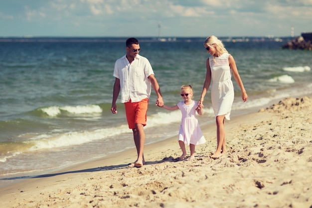 concepto de familia, vacaciones, adopción y personas - hombre feliz, mujer y niña con gafas de sol caminando en la playa de verano