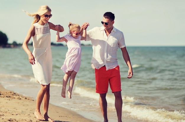 concepto de familia, vacaciones, adopción y personas - hombre feliz, mujer y niña con gafas de sol caminando en la playa de verano