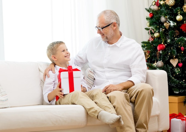 concepto de familia, vacaciones, generación, navidad y personas - abuelo y nieto sonrientes con caja de regalo sentados en el sofá en casa