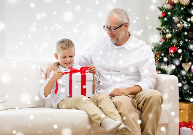 concepto de familia, vacaciones, generación, navidad y personas - abuelo y nieto sonrientes con caja de regalo sentados en el sofá en casa