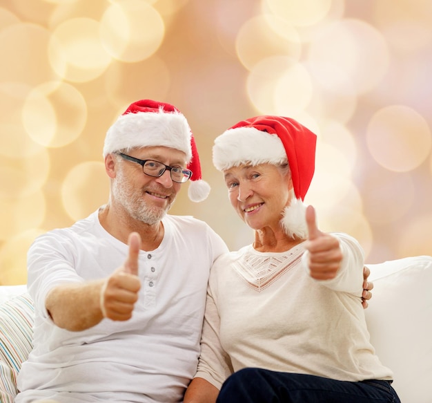 concepto de familia, vacaciones, navidad, edad y personas - feliz pareja de ancianos con sombreros de ayudante de santa sentados en un sofá sobre fondo de luces beige y mostrando un gesto de aprobación