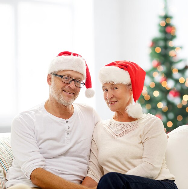 concepto de familia, vacaciones, navidad, edad y personas - feliz pareja de ancianos con sombreros de ayudante de santa sentados en un sofá sobre la sala de estar y el fondo del árbol de navidad