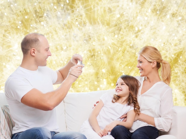concepto de familia, vacaciones, tecnología y personas - madre, padre y niña sonrientes con cámara sobre fondo de luces amarillas
