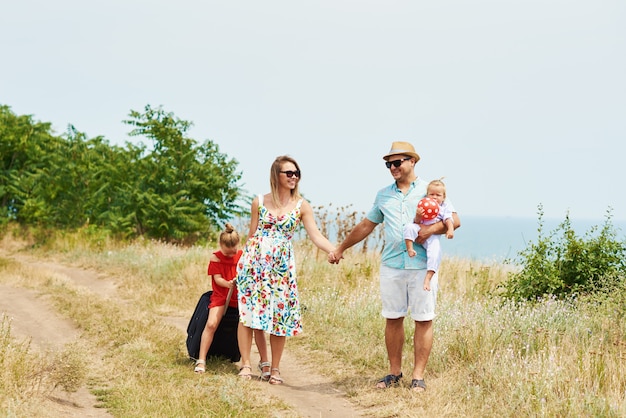 Concepto de familia, vacaciones de verano, adopción y personas: hombre feliz, mujer e hijas en gafas de sol, con maletas divirtiéndose sobre el cielo azul