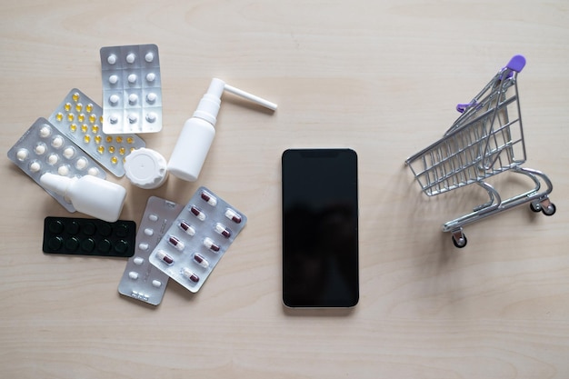 Concepto de farmacia en línea Compra de medicamentos en línea por teléfono Entrega de productos farmacéuticos Mini carro con diferentes tabletas rocía cápsulas y un teléfono inteligente con una pantalla negra en blanco