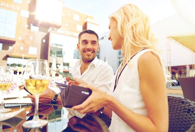 concepto de fecha, personas, pago y finanzas - pareja feliz con billetera y copas de vino pagando la cuenta en el restaurante