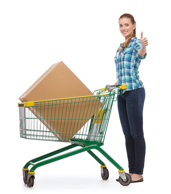 concepto de felicidad, compras y personas - mujer joven sonriente con carrito de compras y caja grande mostrando los pulgares hacia arriba