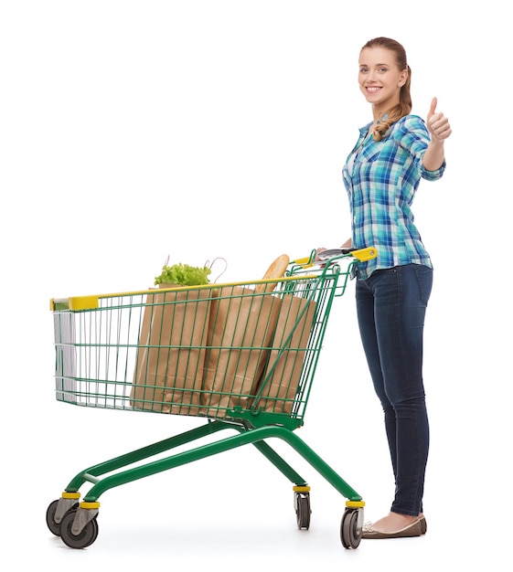 concepto de felicidad, compras y personas - mujer joven sonriente con carrito de compras y comida mostrando los pulgares hacia arriba