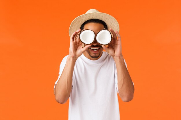 Concepto de felicidad, vacaciones y turismo. Divertido chico afroamericano despreocupado con camiseta blanca, sombrero de verano, sosteniendo cocos en los ojos y sonriendo divertido, bromeando, de pie naranja