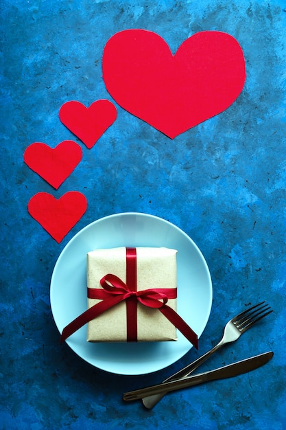 Concepto festivo. regalo en papel ecológico artesanal con cinta roja en placa azul con un tenedor y cuchillo sobre una mesa azul con corazones. cumpleaños, día de san valentín u otros saludos universales