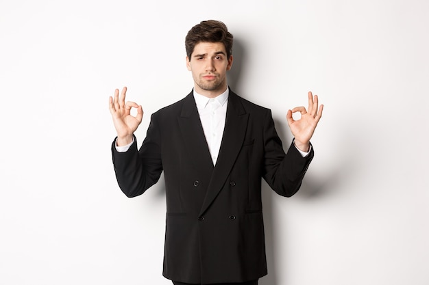 Concepto de fiesta de año nuevo, celebración y estilo de vida. Retrato de hombre guapo seguro en traje negro, mostrando signo bien y aprobar algo, de pie sobre fondo blanco.